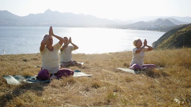 Group of senior people doing yoga exercises at the mountain. Elderly man and two woman sitting on the dry grass meadow at sunny day while making asans against sea and mountains on horizon