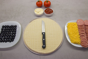 pizza preparation process top view: two round blanks of pizza dough with salami olives, cheese on plates with two tomatoes and two sauces next to it on a gray table with texture