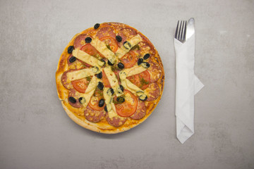 round baked pizza with salami and olives and cheese with a fork and knife on the sides close-up top view