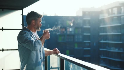 Handsome caucasian middle aged man stands in the balcony with great views and drinks hot coffee or tea, sun shine, enjoying beautiful morning feeling happy - Powered by Adobe