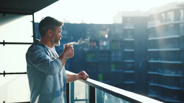 Handsome caucasian middle aged man stands in the balcony with great views and drinks hot coffee or tea, sun shine, enjoying beautiful morning feeling happy
