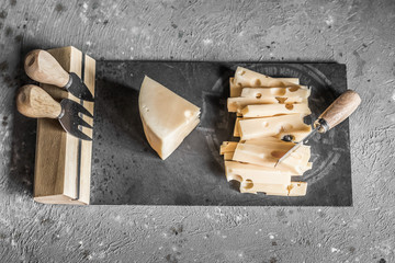 Slice of hard cheese on a black board with cheese knives. Gray concrete background. Top view.