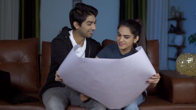 Happy newly married pair discussing the interiors of their new house - future planning concept. Cute Indian couple happily planning for their new home while looking at the blueprints or 3D drawings...