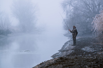 Obraz premium Fisher man fishing with spinning rod on a river bank at misty foggy winter, spin fishing, prey fishing