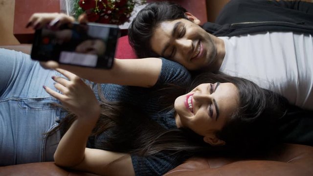 Cute boyfriend girlfriend clicking selfies with wide toothy smiles on valentine's day. Top view shot of a beautiful young couple taking pictures with a smartphone while lying together on a couch - ...