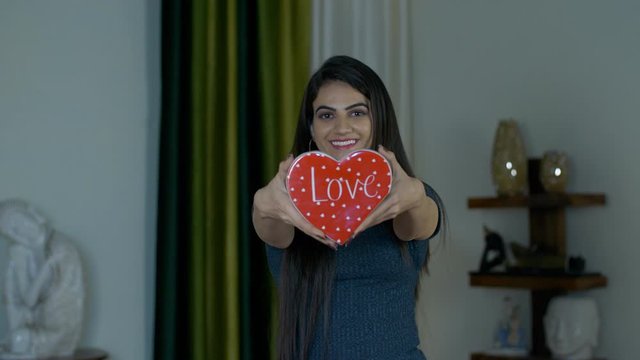 Pretty Indian girl smiling and showing a red heart symbol of love to the camera. Young happy female holding a ceramic heart-shaped toy with a wide toothy smile on her face on Valentine's day celebr...