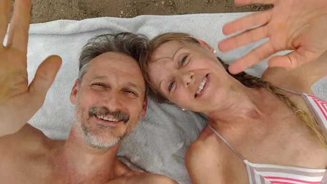 Portrait of man and happy woman, waving the selfie camera of the smarphone on the beach. Background towel. Greetings with smart phone