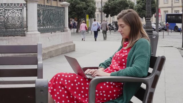Young woman freelancer, student in red dress is working at laptop, sitting on a bench in the city square, enjoying sunny warm weather and her lifestyle, Slow motion.
