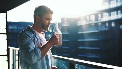 Dreaming european middle aged man stands in the balcony with great views and drinks hot coffee or tea, sun shine, enjoying beautiful morning feeling happy slow motion - Powered by Adobe