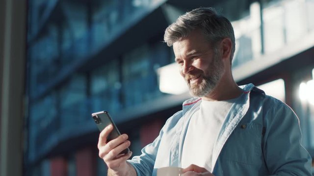 Close up view of happy mature man uses phone drinks coffee good news wonderful emotions wins in the modern balcony texting scrolling tapping great morning technology communication sunny day