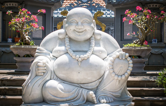 A statue of a seated and smiling Buddha in the territory of one of the Pagodas located in Hoi An city, Vietnam. The statue is made of white marble.
