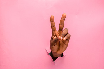 black hand through a hole in a cardboard making v sign with two fingers, pink background