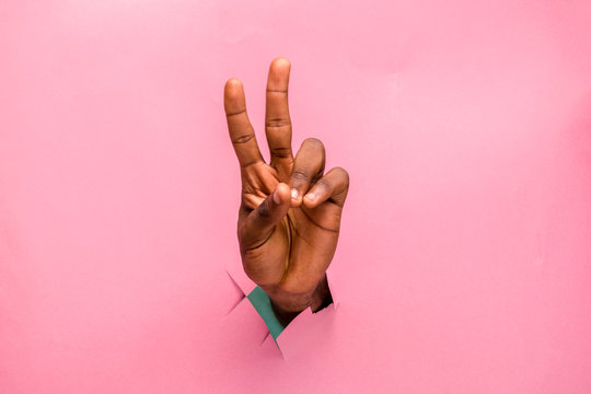 Black Person Hand Through A Hole In A Cardboard Making V Sign With Two Fingers, Pink Background