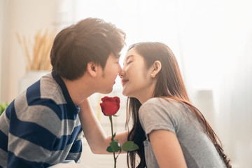 Obraz na płótnie Canvas Close up shot Asian pretty woman and handsome man holding rose flower together sitting on bed in bedroom, closing eyes, smile and kissing with romantic moment. Couple lover and lifestyle in Valentine.