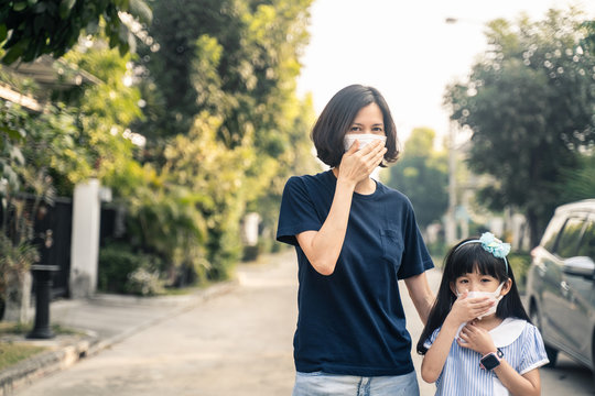 Asian woman and little young daughter wearing mask for prevent dusk pm 2.5 bad air pollution. Mother and girl kid holding mask and having a cough due to bad smell and allergic symptom stand on street.