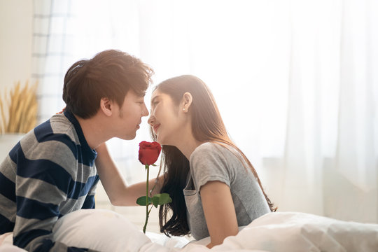 Asian pretty woman and handsome man holding rose flower together sitting on bed in bedroom, closing eyes, smile and kissing with romantic moment. Couple lover and lifestyle in Valentine day concept.