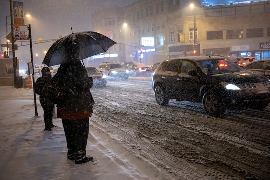 man is standing outside with umbrella in the extreme cold and snow in Chicago area during a blizzard during a winter night in January.  traffic is slowing down due to weather conditions