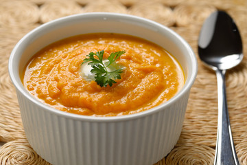 Carrot soup with cream and parsley in white bowl on woven grass placemat