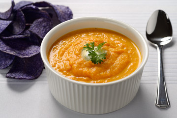 Carrot soup with cream and parsley in white bowl with blue potato chips