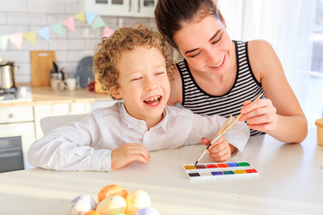 happy young dark-haired mom and her curly blond son paint eggs for easter in the white kitchen in the Scandinavian style © Olga Gimaeva