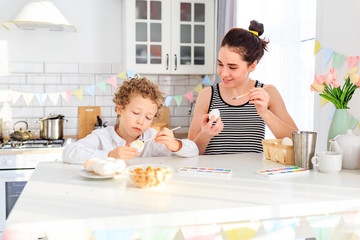 Obraz na płótnie Canvas happy young dark-haired mom and her curly blond son paint eggs for easter in the white kitchen in the Scandinavian style