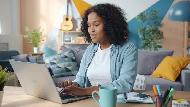 Excited young African American business lady is enjoying success at freelance work at home looking at laptop screen having fun celebrating good news.