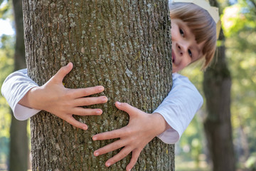 Portrait of a pretty smiling child girl in a paper crown on her head embracing a tree trunk in summer park. Love and care about nature concept.