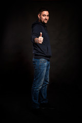 A young man in jeans and a hoodie holds a thumbs up. Full height. Focus in the foreground. Black background. Vertical.
