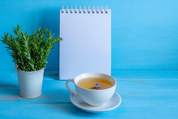 Cup of rosemary tea, rosemary sprigs in miniature basket, blank page of note book on blue wooden background with copy space 
