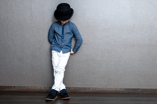 A gentleman boy in a black bowler hat in white jeens stands against the dark wall. Copy space.