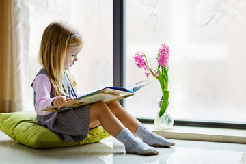 cute little girl with blonde hair sitting on the floor on balcony and reading book. Education,...