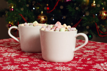 Obraz na płótnie Canvas White mugs of hot chocolate beverage with colorful marshmallow on red cloth table with snowflakes on Christmas tree background 