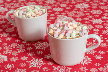 White mugs of hot chocolate beverage with colorful marshmallow on red cloth table with snowflakes 