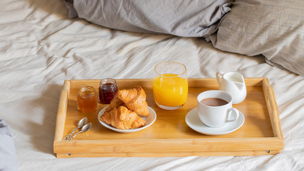 Fototapeta na wymiar Breakfast on the bed inside a bedroom. Cup of coffee, orange juice, croissant and jam on wooden try. Good morning concept.