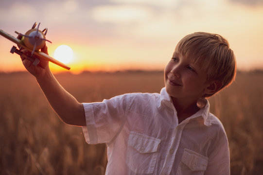 Happy Child Playing With A Toy Plane In Nature During Summer Sunset. Boy In A  White Shirt With A Plane In Hands On Wheat Field. Kid Holds A Wooden Airplane And Dreams Of Being A Pilot, On The Nature