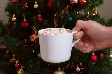 Male hand holding white mug with hot chocolate beverage with colorful marshmallow on Christmas tree background 