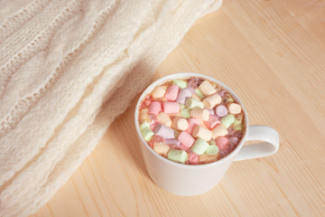 Obraz na płótnie Canvas Hot chocolate beverage with colorful marshmallows on light wooden background with white fluffy knitting blanket. Hygge style coziness concept 
