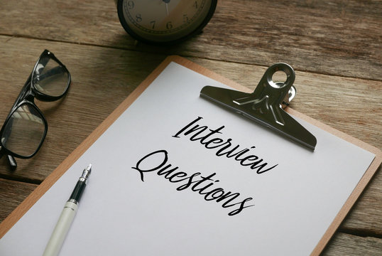 Glasses,clock,pen and clipboard with paper written Interview Questions on wooden background.