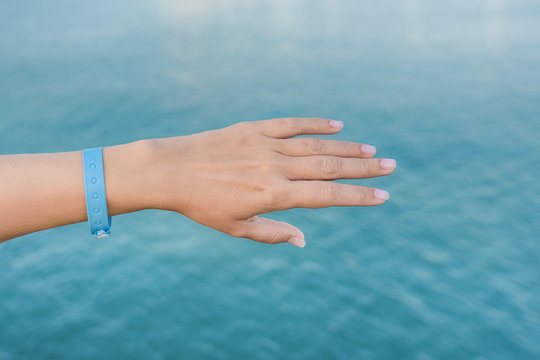 Closeup view of beautiful white female hand wearing blue rubber wristband. Hand isolated at blurry sea sunny water bokeh background. Happy travel and all inclusive system of hotel resorts concept.