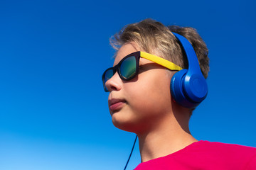 Closeup portrait of cute cool teen boy standing outdoors isolated on sunny clear blue sky background. Kid wearing headphones and sunglasses. Horizontal color photography.