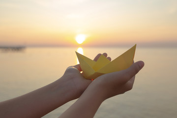 Closeup view of yellow folded paper boat in hands of young kid isolated at sunny golden sunset or sunrise sky and sea landscape background. Happy and safe tourism concept. Horizotal color photography. - obrazy, fototapety, plakaty