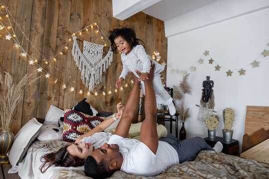 Positive young mixed race family cute young mother African American dad and charming smiling daughter have fun on the bed on Friday evening. Family time concept