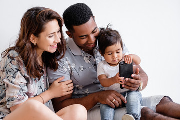 Positive young mixed race family charming mom african-american dad and curious daughter watching photos together on smartphone sitting on floor at home on the white wall. Concept of family and gadgets