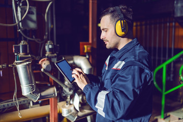 Fototapeta na wymiar Focused handsome factory worker in working clothes and with antiphons using tablet. Factory interior.