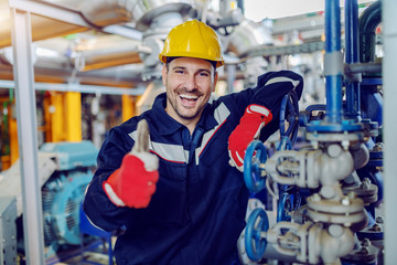Smiling handsome caucasian worker in protective working clothes and with helmet on head leaning on boiler and giving thumbs up while standing in factory.