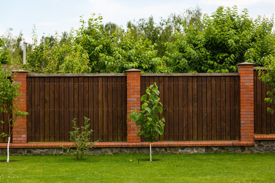 New wooden fence with massive stone brick pillars. Green lawn and trees, daytime