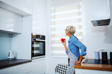 Charming Caucasian blonde senior woman in apron leaning on kitchen counter, drinking wine and waiting for dish to be baked.