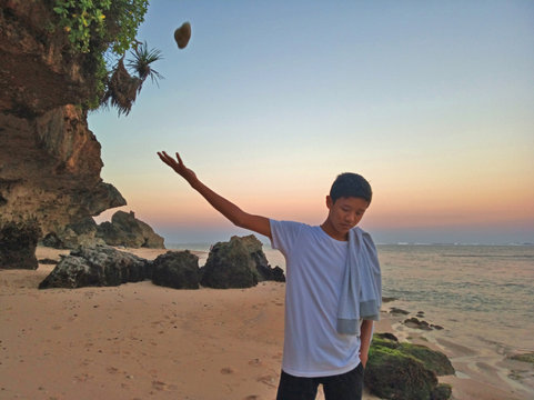 Young man throwing stones in the air on the rocky beach at sunset.