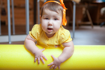 Little girl portrait, baby lying on yellow foam roller in domestic gym, looking at camera