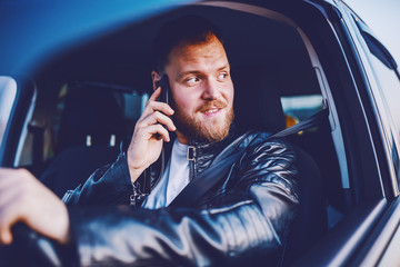 Young handsome caucasian bearded blond man in leather jacket driving his car and using smart phone.